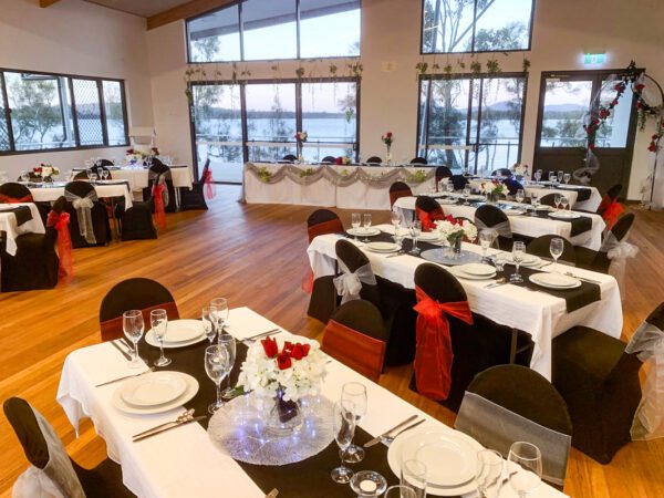 Elegant event venue with decorated tables featuring white tablecloths, black table runners, floral centerpieces, and chairs adorned with red and silver sashes, overlooking scenic outdoor views through large windows.
